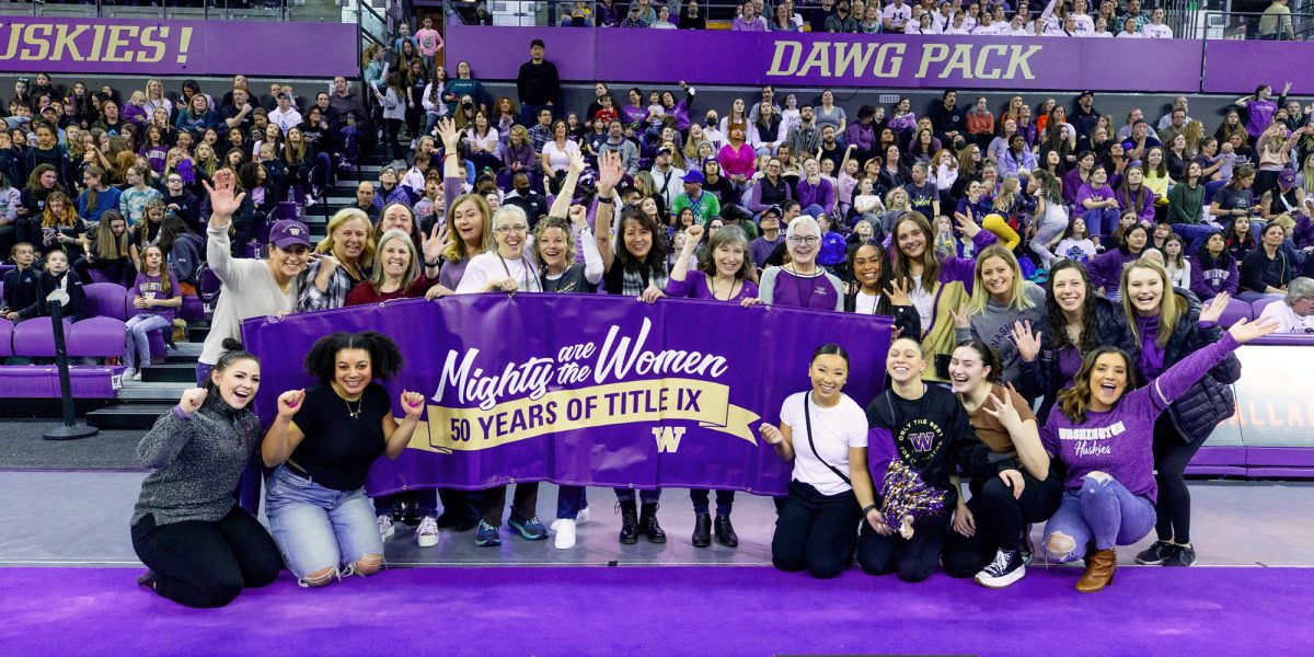 Generations of UW Gym Dawgs hold Mighty are the Women Banner in front of crowd