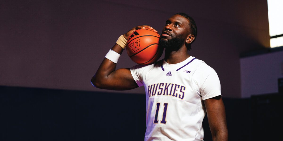 Frank Kepnang holds basketball on shoulder and looks off into the distance in a dark room with one bright window