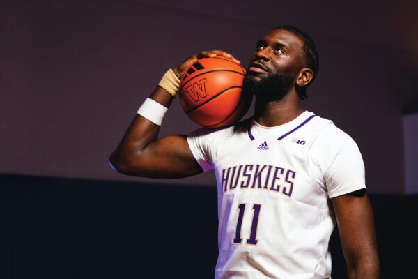 Frank Kepnang holds basketball on shoulder and looks off into the distance in a dark room with one bright window